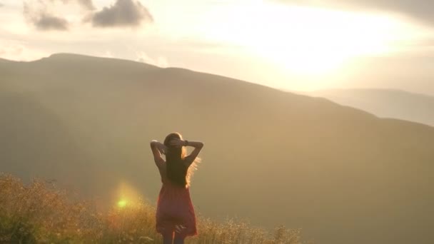 Joven mujer feliz viajero en vestido rojo de pie en la ladera cubierta de hierba en una noche ventosa en las montañas de verano disfrutando de la vista de la naturaleza al atardecer. - Imágenes, Vídeo
