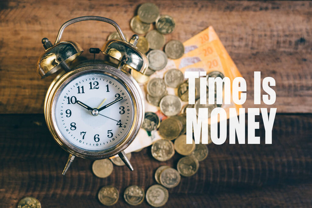 Время - это денежная концепция - золотые колокольные часы, деньги и монеты на фоне деревянного стола со словом "время - золото" - Фото, изображение