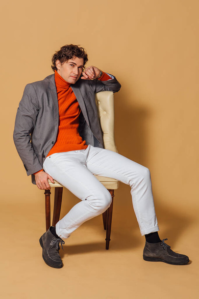 Fashion portrait of a tall handsome man dressed in orange turtleneck, white jeans and grey jacket sitting on an armchair on the beige background - Foto, afbeelding