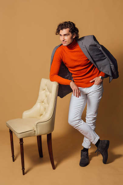 Fashion portrait of a tall handsome man dressed in orange turtleneck, white jeans and grey jacket posing with an armchair on the beige background - 写真・画像