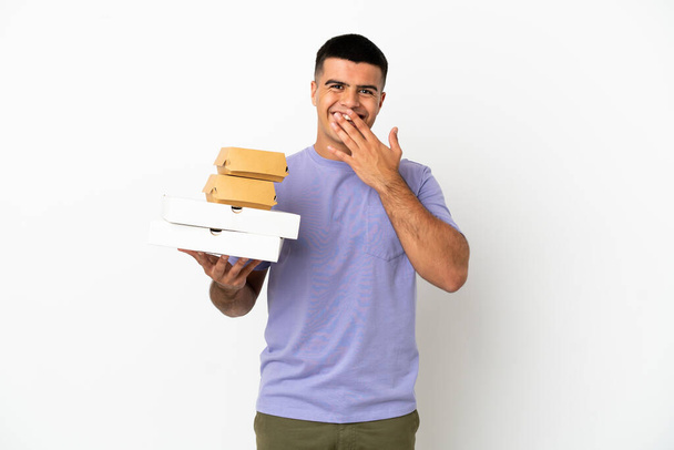 Νεαρός όμορφος άντρας που κρατάει πίτσες και μπιφτέκια πάνω από απομονωμένο λευκό φόντο χαρούμενος και χαμογελαστός που καλύπτει το στόμα με το χέρι - Φωτογραφία, εικόνα