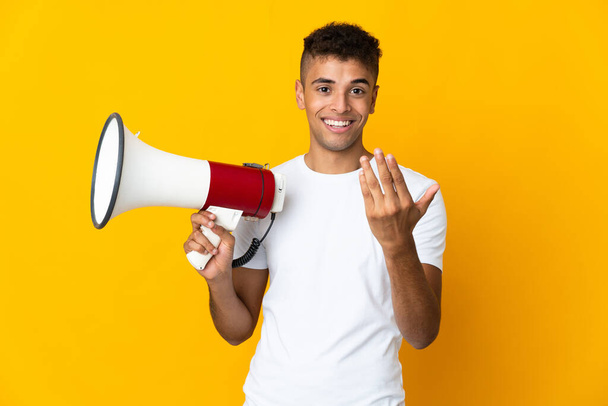 Young brazilian man isolated on yellow background holding a megaphone and inviting to come with hand - Photo, Image