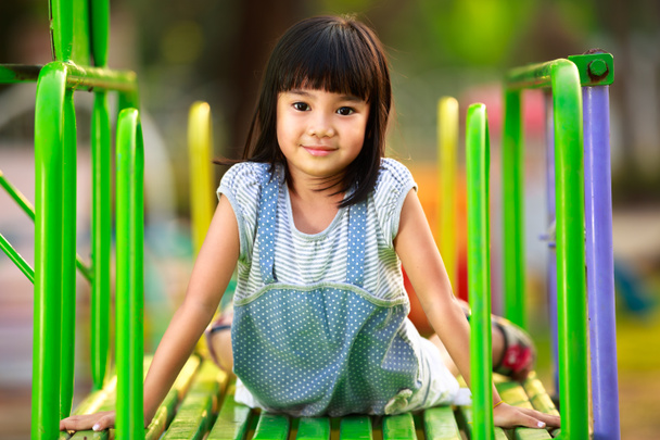 Маленькая азиатская девочка, сидящая на горке на детской площадке
 - Фото, изображение