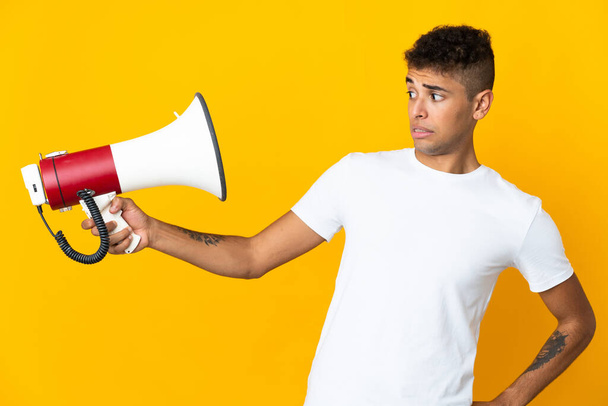 Young brazilian man isolated on yellow background holding a megaphone with stressed expression - Photo, Image