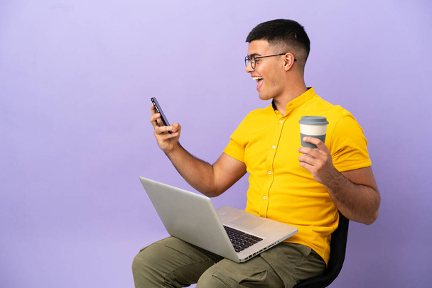 Jeune homme assis sur une chaise avec ordinateur portable tenant café à emporter et un mobile - Photo, image