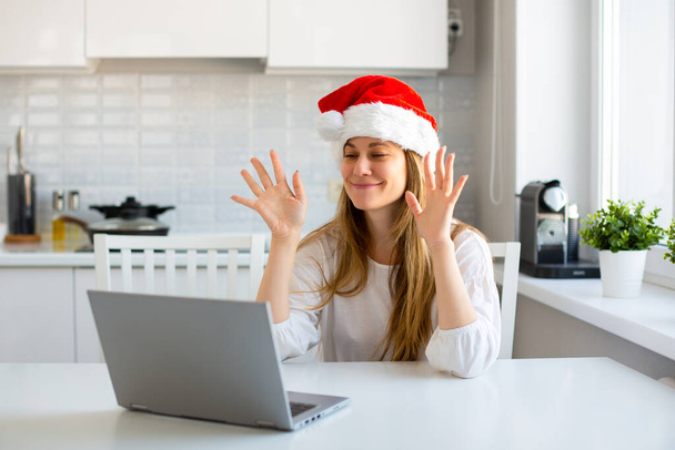 Portrait of a woman in Santa hat in front of laptop monitor. She celebrating Christmas during quarantine - online meeting with friends and relatives. - Photo, Image