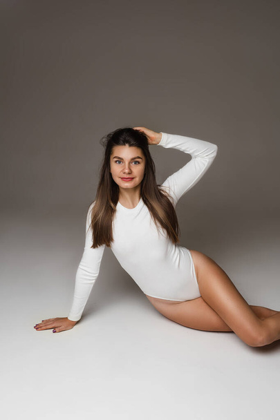 Beautiful young fit girl in white bodysuit, tanned slim female model with long healthy hairs posing on studio floor - Фото, зображення