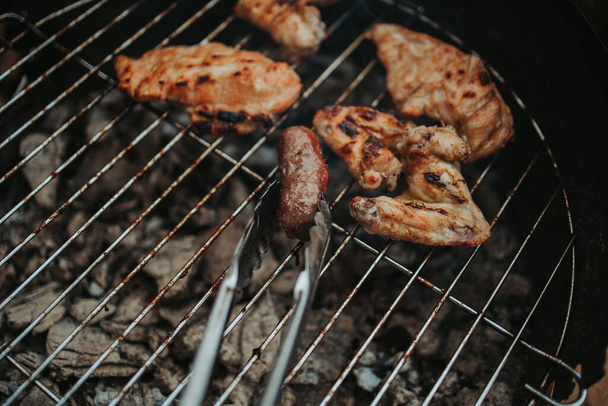 A pair of tongs holding a kebab over a grill with chicken wings and breasts cooking on it in the background - Fotó, kép