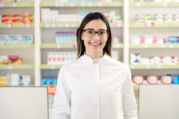 Egy gyógyszerész portréja egy gyógyszertárban. Egy közeli kép egy egyenruhás gyógyszerész nőről, szemüveggel a gyógyszertár közepén, egy gyógyszeres polc előtt. Boldogan elvégzem a munkát. - Fotó, kép