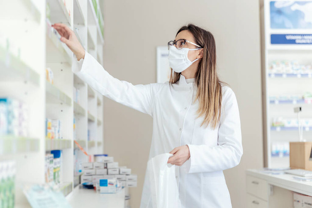 Trabajo en una farmacia. Embalaje de medicamentos en una bolsa. Mujer empleada como farmacéutica y vestida con un uniforme blanco con una máscara protectora en la cara en bolsas que empaquetan cajas de medicamentos y suplementos - Foto, imagen