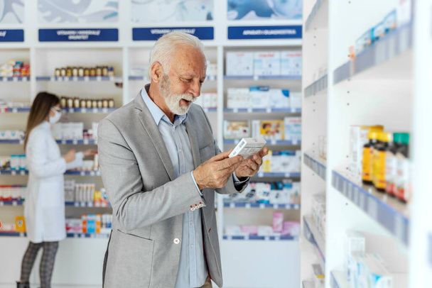 Gyógyszerek vásárlása gyógyszertárakban és gyógyszertárakban. Közelkép egy elegáns, érett férfiról, védőmaszkkal az arcán, amint egy gyógyszeres dobozból és kiegészítőkből álló nyilatkozatot olvas fel. - Fotó, kép