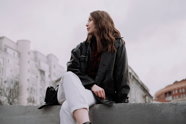 Hymyilevä nuori nainen mustassa nahkatakissa ja valkoisissa farkuissa istumassa betonilla, katsoen vasemmalle. Kaupunkityyliä ja katumuotia. Tyttö, jolla on kihara kampaus rento asu. Profiilikuva - Valokuva, kuva