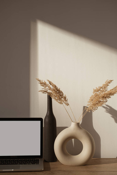 Αισθητικός χώρος εργασίας γραφείου με σκιές ηλιακού φωτός στον τοίχο. Κενό φορητός υπολογιστής οθόνης με το διάστημα αντιγράφων. Γυαλιά, πάμπας γρασίδι σε κομψό βάζο σε ξύλινο τραπέζι. Influencer lifestyle blog - Φωτογραφία, εικόνα