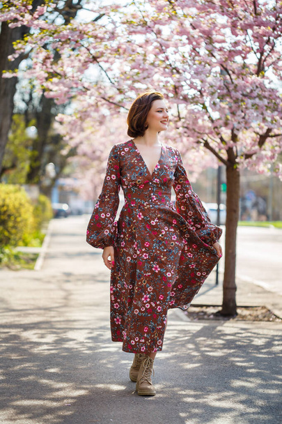 Hermosa mujer joven en un vestido con árboles de sakura en flor. Hace un cálido día soleado de primavera afuera. - Foto, imagen