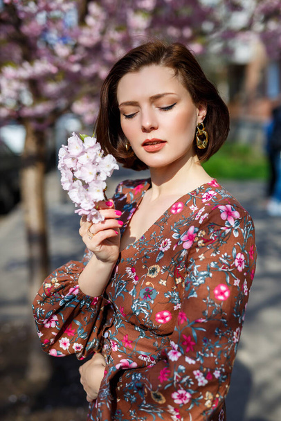 Πορτρέτο ενός χαριτωμένο και όμορφο κορίτσι γυναίκα σε ένα φόρεμα στον κήπο στη μέση της ανθοφορίας sakura. 'νοιξη και ήλιος. Θολή φόντο και επιλεγμένη εστίαση. - Φωτογραφία, εικόνα