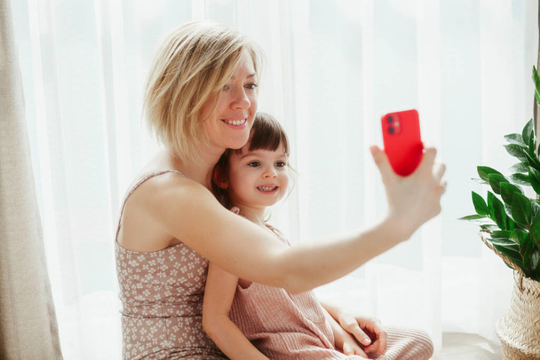 Young woman and her pretty little daughter are sitting together and hugging, taking selfies on a smart phone - Foto, immagini