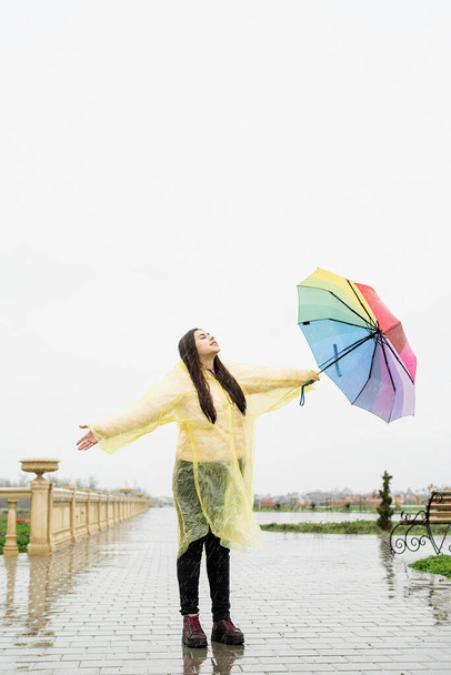 美しいですブルネット女性で黄色レインコート保持虹の傘アウトで雨 - 写真・画像