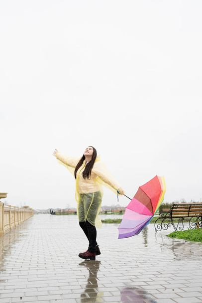 Beautiful brunette woman in yellow raincoat holding rainbow umbrella out in the rain - Photo, Image