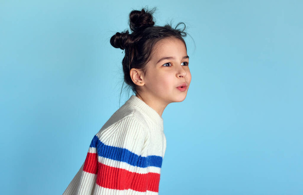 Portrait of a positive little girl with funny hairstyle, blowing, isolated over studio blue color background. Joyful kid has a happy expression on her face. - Foto, imagen
