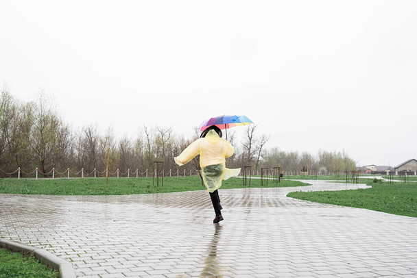 Kobieta w żółtym tęczowym płaszczu trzymająca tęczowy parasol w deszczu. Ludzie z tyłu, widok z tyłu - Zdjęcie, obraz