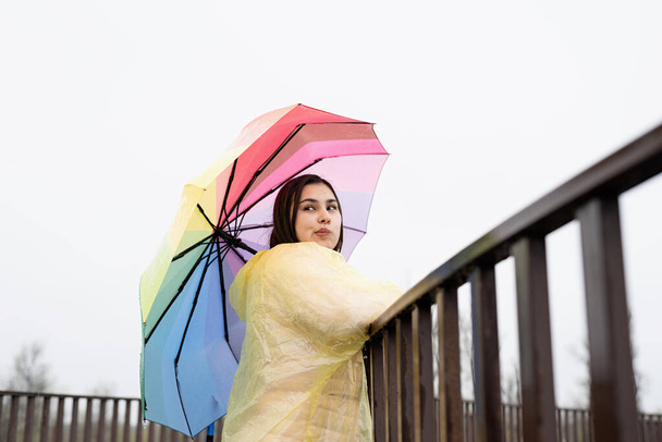 Schöne lächelnde brünette Frau in gelbem Regenmantel mit Regenbogenschirm im Regen, wegschauend. Kopierraum - Foto, Bild