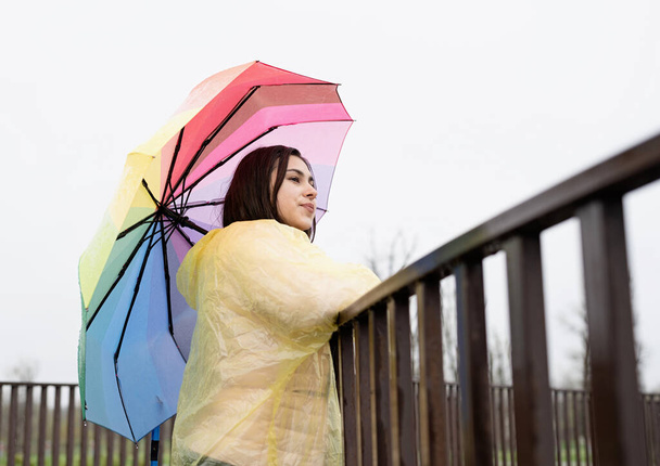 Sarı yağmurluklu güzel, gülümseyen esmer kadın yağmurda gökkuşağı şemsiyesi tutuyor, başka tarafa bakıyor. Boşluğu kopyala - Fotoğraf, Görsel