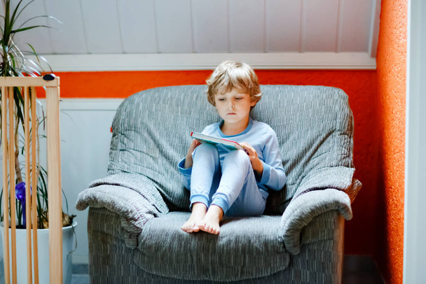 Roztomilý blonďatý kluk z mateřské školky v pyžamu, co čte knížky pro děti v domácím pokoji. Nadšené dítě hlasitě čte, sedí na starém velkém křesle. Školačka, rodina, vzdělání - Fotografie, Obrázek