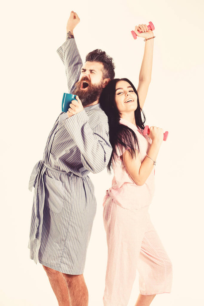 Couple in love in pajama, bathrobe stand back to back. Girl with dumbbell, man with coffee cup, isolated on white background. Morning energy concept. Couple, family on sleepy faces, stretching - Zdjęcie, obraz