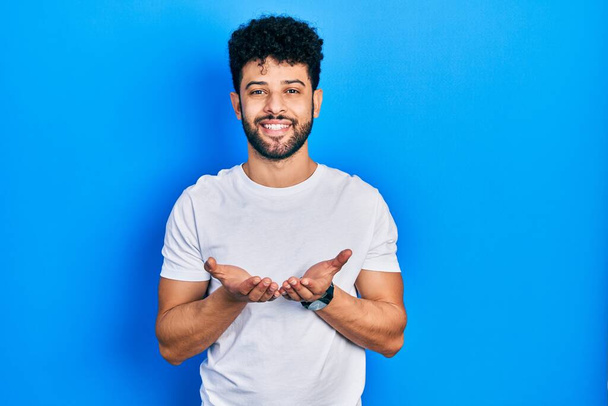 Jeune homme arabe barbu portant un t-shirt blanc décontracté souriant avec les mains paumes ensemble recevant ou donnant geste. maintien et protection  - Photo, image