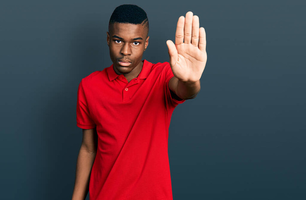 Joven afroamericano vestido con camiseta roja casual haciendo parar de cantar con la palma de la mano. expresión de advertencia con gesto negativo y grave en la cara.  - Foto, imagen