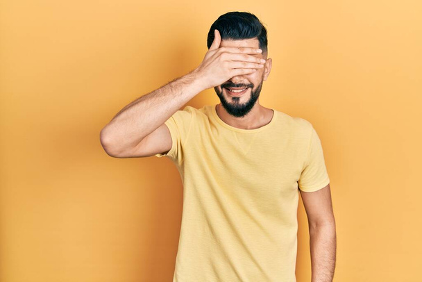 Handsome man with beard wearing casual yellow t shirt smiling and laughing with hand on face covering eyes for surprise. blind concept.  - Photo, Image