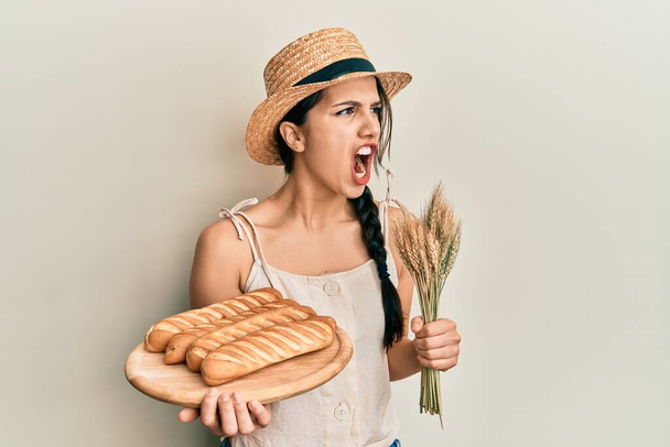 若いヒスパニック系の女性が自家製のパンを保持し、怒っている小麦スパイクと怒りを叫び、怒りで叫んで怒っている。怒りと攻撃的な考え方.  - 写真・画像