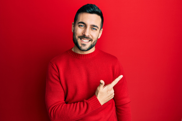 Junger hispanischer Mann in lässiger Kleidung lächelt fröhlich und zeigt mit der Hand und dem Finger zur Seite  - Foto, Bild