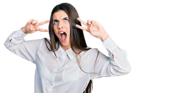 Young brunette teenager doing peace gesture close to eyes angry and mad screaming frustrated and furious, shouting with anger looking up.  - Photo, Image