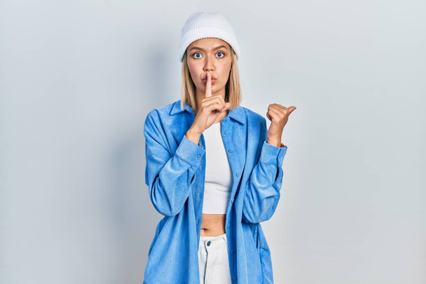 Beautiful blonde woman wearing wool hat asking to be quiet with finger on lips pointing with hand to the side. silence and secret concept.  - Photo, Image