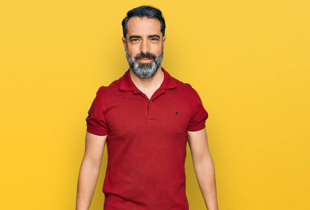 Мужчина средних лет с бородой в обычной красной футболке выглядит позитивно и счастливо стоя и улыбаясь с уверенной улыбкой, показывая зубы  - Фото, изображение