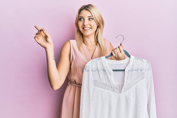 Young caucasian woman holding hanger with t shirt smiling happy pointing with hand and finger to the side  - Foto, Imagem