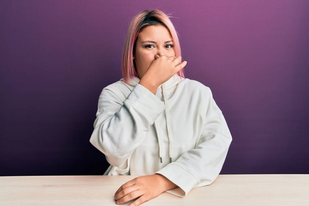 Latijns-Amerikaanse vrouw met roze haar in casual sweatshirt zittend op de tafel ruiken iets stinkend en walgelijk, ondraaglijk geur, adem inhouden met vingers op de neus. slechte geur  - Foto, afbeelding
