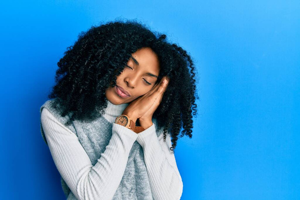 Afroamerikanerin mit Afrohaaren trägt lässigen Winterpullover, schläft müde träumend und posiert mit den Händen zusammen, während sie mit geschlossenen Augen lächelt.  - Foto, Bild