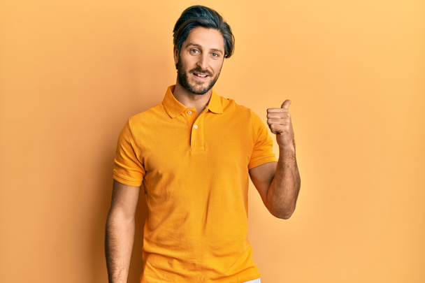 Fiatal spanyol férfi alkalmi sárga pólóban mosolyog boldog arccal, és feltartott hüvelykujjal oldalra mutat..  - Fotó, kép