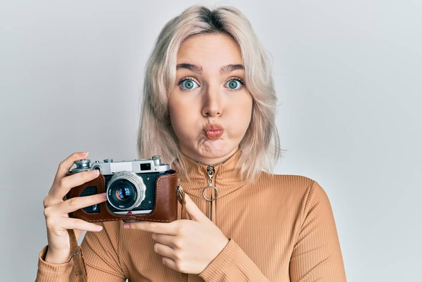 Νεαρό ξανθό κορίτσι κρατώντας vintage κάμερα puffing μάγουλα με αστείο πρόσωπο. στόμα φουσκωμένο με αέρα, που πιάνει αέρα.  - Φωτογραφία, εικόνα