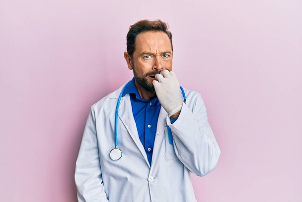 Keski-ikäinen mies yllään lääkäri yhtenäinen ja stetoskooppi näköinen stressaantunut ja hermostunut kädet suulla puree kynnet. ahdistuneisuusongelma.  - Valokuva, kuva