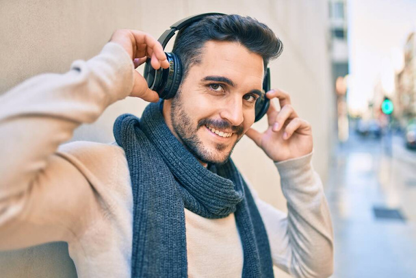 Νεαρός Ισπανός χαμογελάει χαρούμενος ακούγοντας μουσική με ακουστικά στην πόλη. - Φωτογραφία, εικόνα