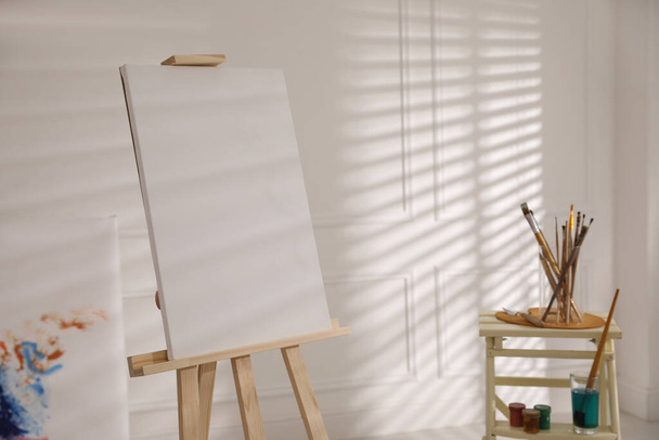 Wooden easel with empty canvas and brushes in art studio - Fotoğraf, Görsel