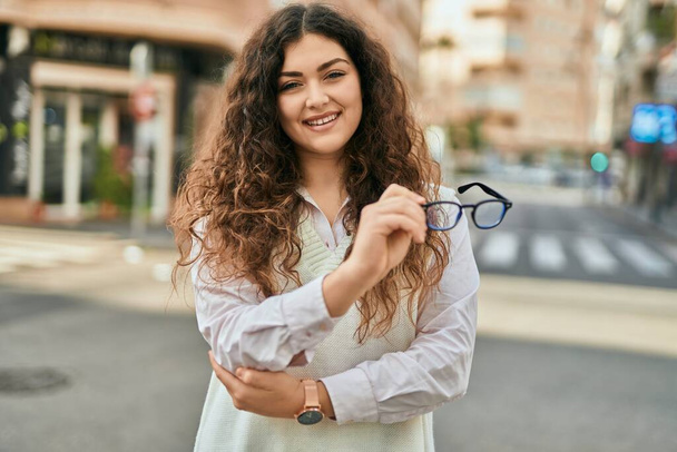 Jeune femme hispanique souriant heureux tenant des lunettes à la ville. - Photo, image