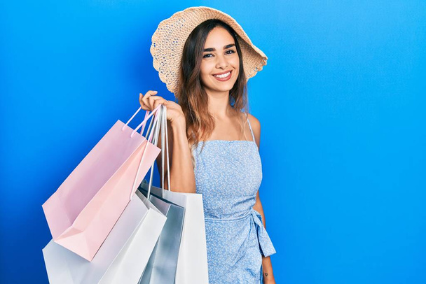 Joven chica hispana sosteniendo bolsas de compras con aspecto positivo y feliz de pie y sonriendo con una sonrisa confiada mostrando los dientes  - Foto, imagen
