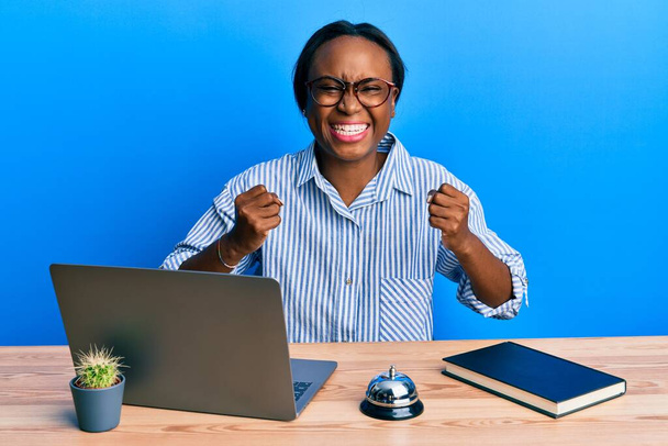 Jonge Afrikaanse vrouw werkt bij de receptie van het hotel met behulp van laptop enthousiast voor succes met opgeheven armen en ogen gesloten vieren overwinning glimlachen. winnaarsconcept.  - Foto, afbeelding