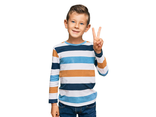 Αξιολάτρευτο καυκάσιο παιδί που φοράει καθημερινά ρούχα χαμογελώντας με χαρούμενο πρόσωπο να κλείνει το μάτι στην κάμερα κάνοντας το σήμα της νίκης. Νούμερο δύο..  - Φωτογραφία, εικόνα
