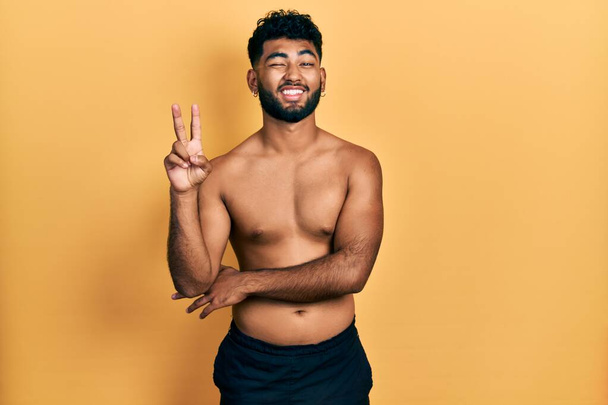 Arabische man met baard draagt zwemkleding zonder shirt glimlachend met een vrolijk gezicht knipogend naar de camera terwijl hij overwinningstekens doet. nummer twee.  - Foto, afbeelding