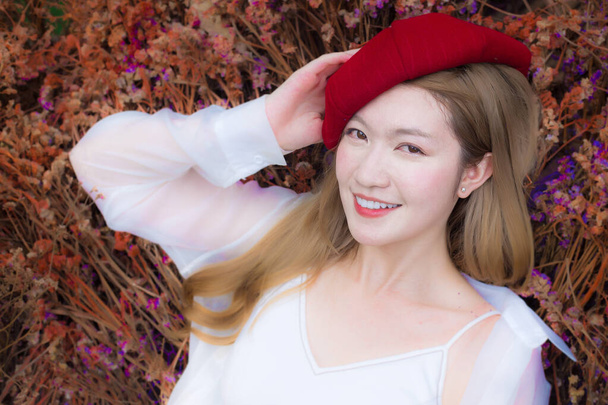 Portrait Asian beautiful lady who wears white dress and red cap smiles in colored dried flowers as natural background. - 写真・画像